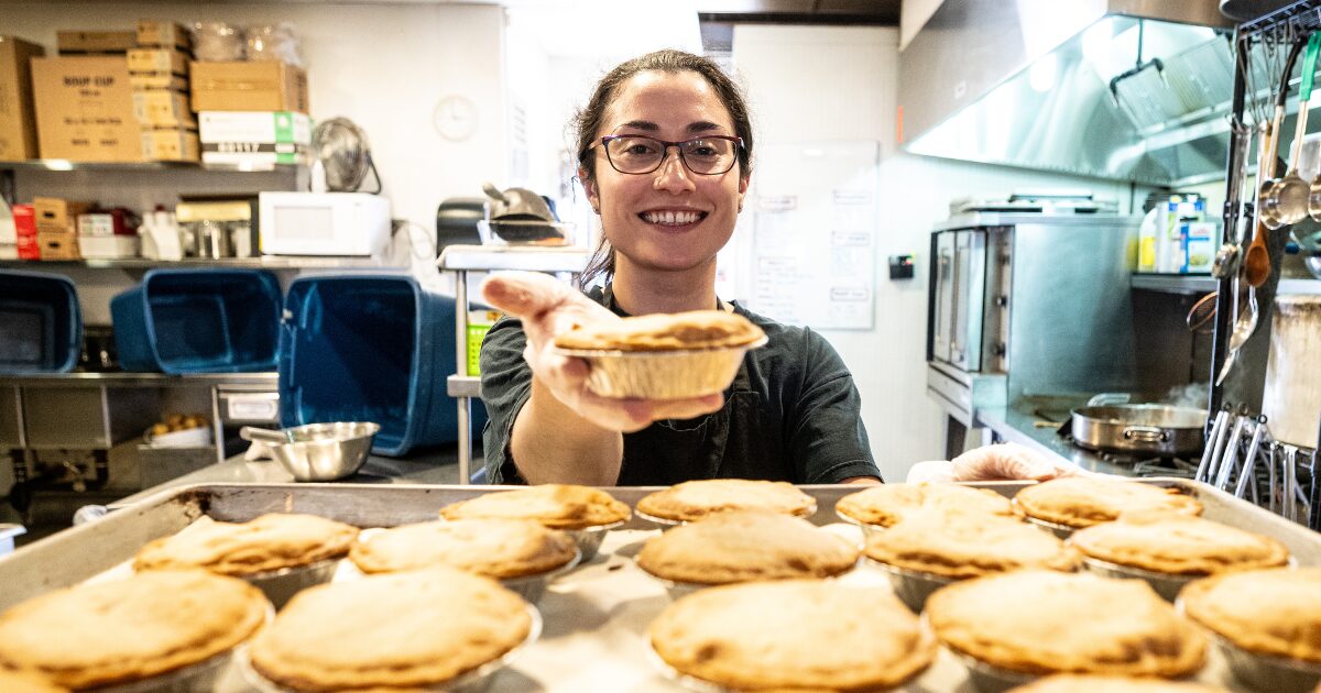 This photograph from the WISH Kitchen show a volunteer with dark hair and glasses handing a freshly baked pie to the camera. There is a tray with 12 other pies in front of her, and lots of kitchen equipment in the background.